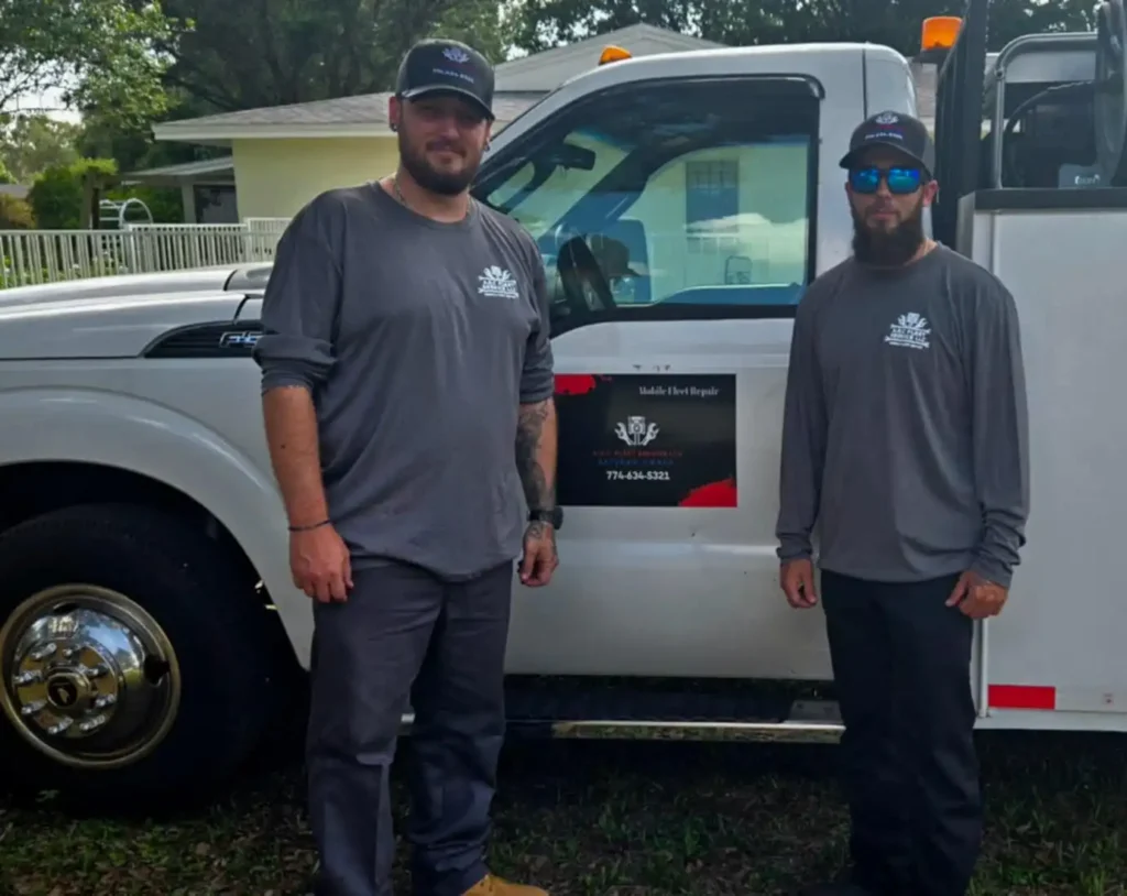 Two men in matching gray shirts and hats stand in front of a white work truck with a business logo and phone number, ready to help as your local mobile diesel mechanic.